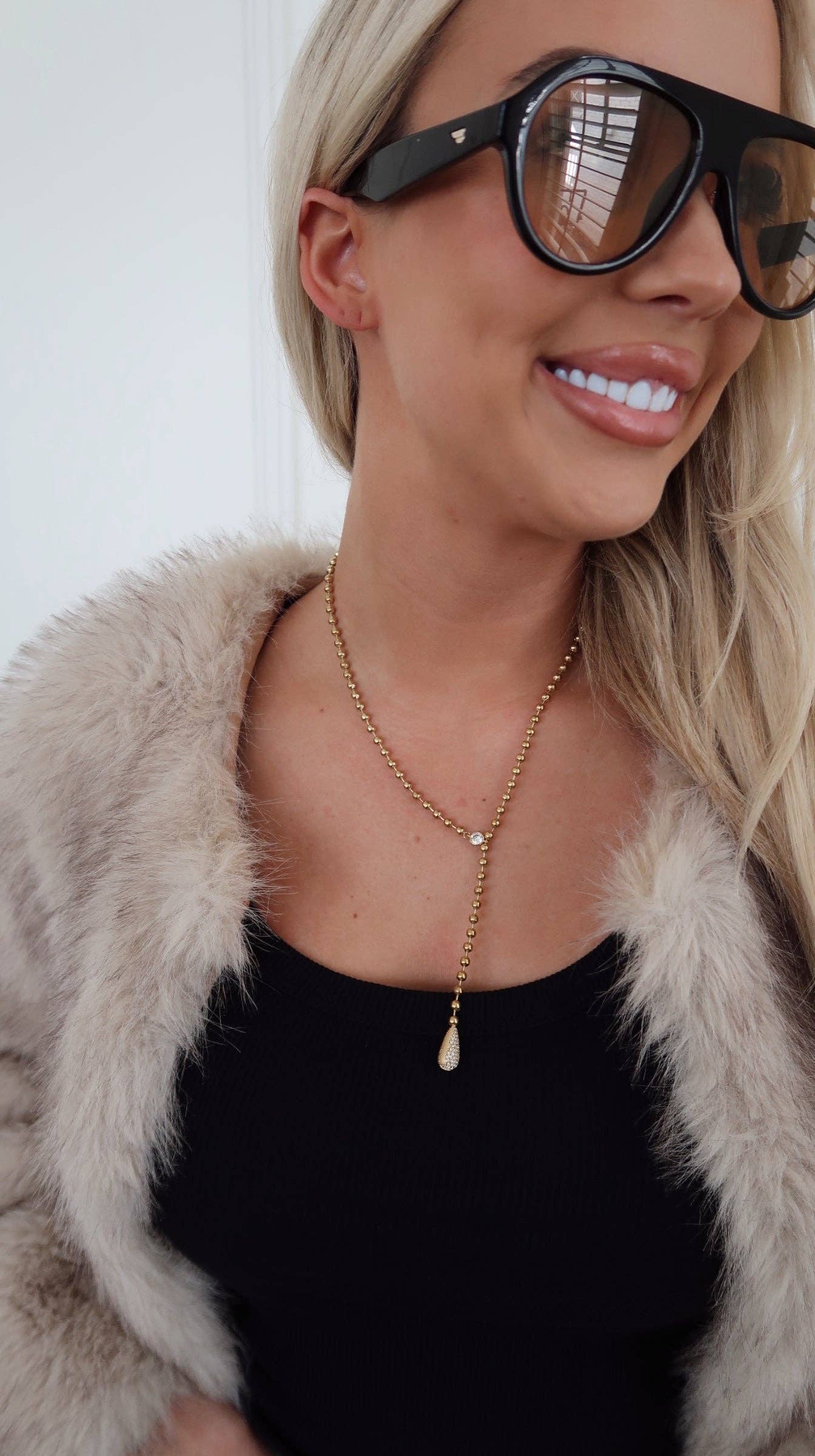 The Lany Necklace: gold beaded chain with teardrop pendant, adjustable 16-18 inches, worn by a woman in sunglasses.